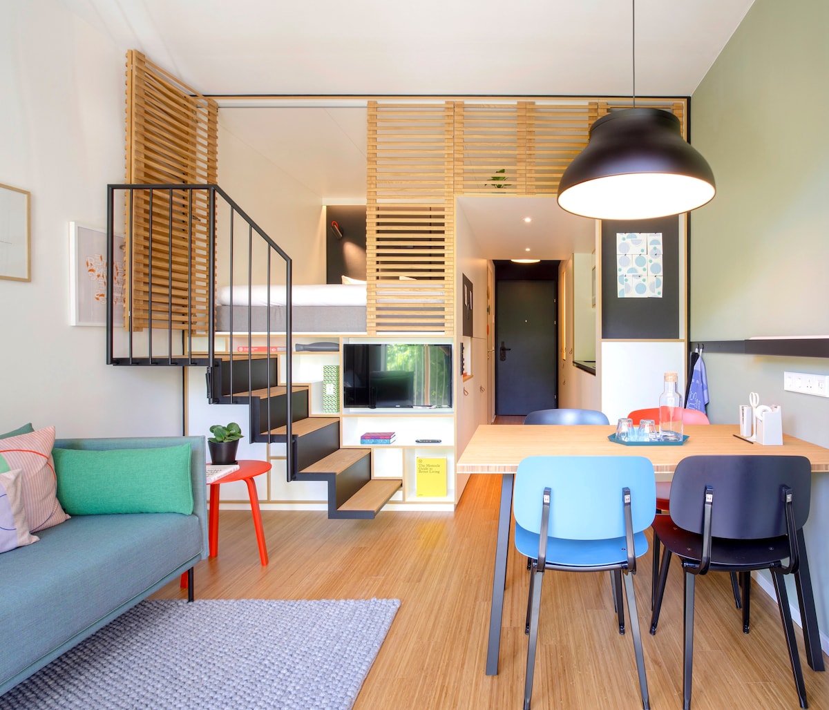 A contemporary living space features a green sofa and a dining area with a wooden table and black chairs. A staircase with open shelving leads to a lofted sleeping area. Natural light fills the room, showcasing the minimalist design elements and warm wood tones.