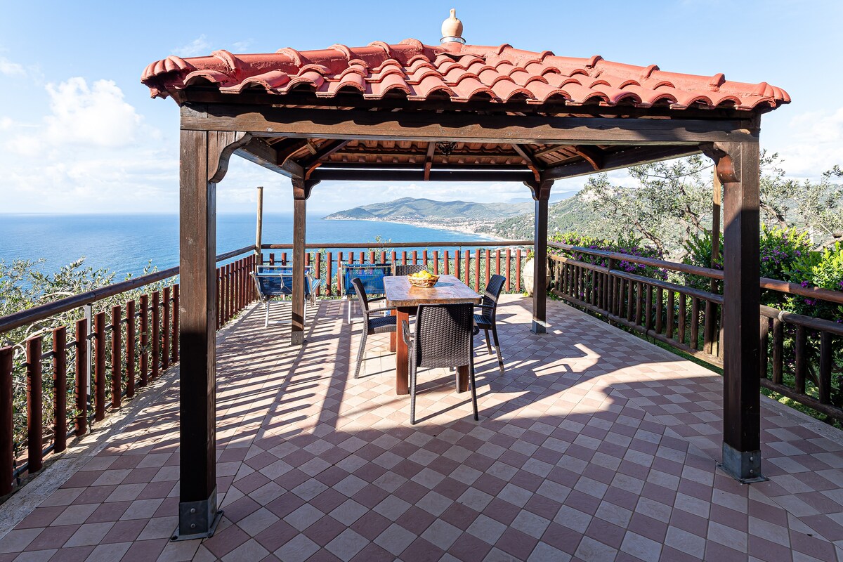 A spacious covered patio is showcased, featuring a tiled floor and a wooden structure with a terracotta roof. Outdoor dining furniture, including a table and chairs, is positioned to provide unobstructed views of the surrounding hillside and the shimmering sea in the distance.