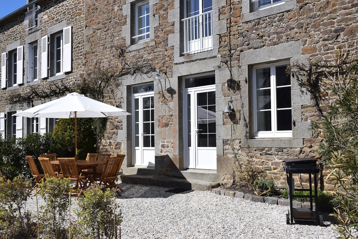 An outdoor seating area is visible, featuring a wooden dining table surrounded by matching chairs. A white umbrella provides shade over the table, and a grill stands nearby on a gravel surface. The stone facade of the building adds rustic charm.