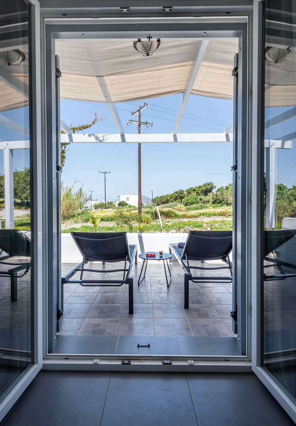 The view from the patio features a light-filled entry with doors open to a spacious outdoor area. Two lounge chairs and a small table are positioned under a shade structure, surrounded by greenery and an unobstructed sky.