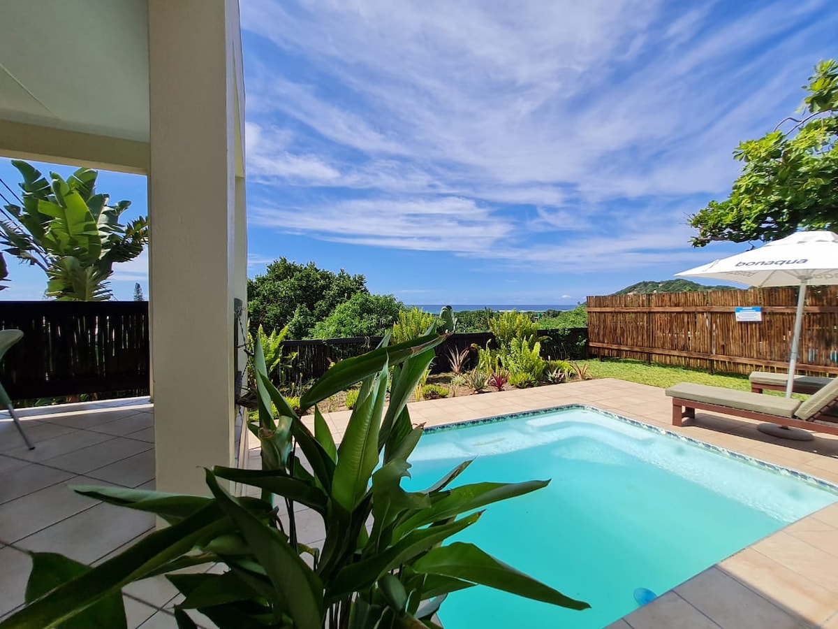 The outdoor patio features a private splash pool surrounded by lush greenery, offering views of the ocean and town. A sun umbrella and lounge chairs are positioned for relaxation, while a wooden fence provides privacy. The sky is clear with wispy clouds overhead.