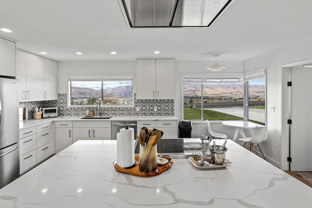 A modern kitchen is presented with a spacious island and white cabinetry. Large windows provide views of the landscape, while patterned backsplash and stainless steel appliances enhance the contemporary design. Natural light brightens the area, creating a welcoming space for cooking and gathering.