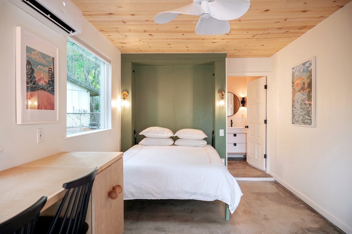 A tastefully remodeled guest suite features a spacious bed with neatly arranged pillows against a green accent wall. Natural light fills the room through windows, complemented by wood paneling on the ceiling. A small desk with black chairs is present, and a doorway leads to a bathroom.