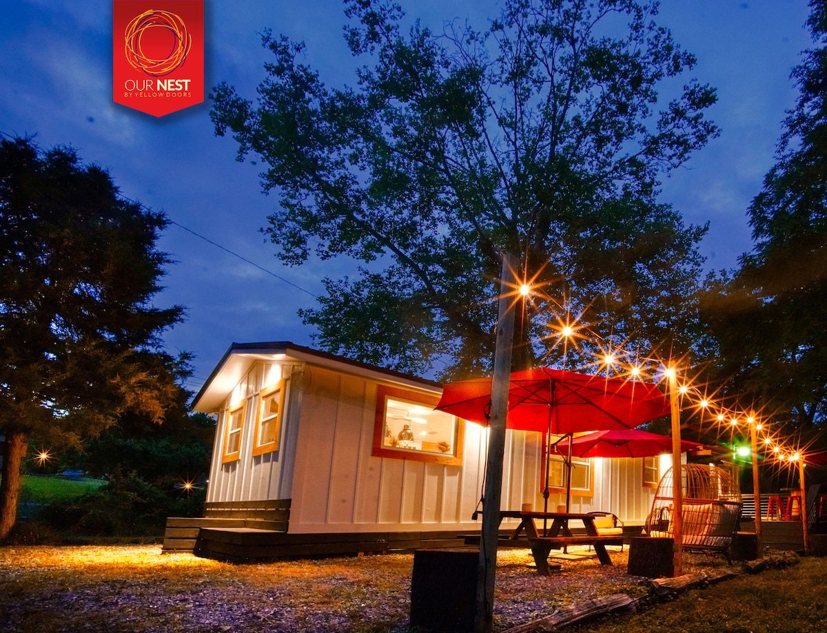 The exterior of the mobile cabin is highlighted by warm outdoor lighting, creating a welcoming ambiance. A picnic table with an umbrella is set nearby on gravel, surrounded by trees, while the cabin features illuminated windows showcasing a cozy interior.