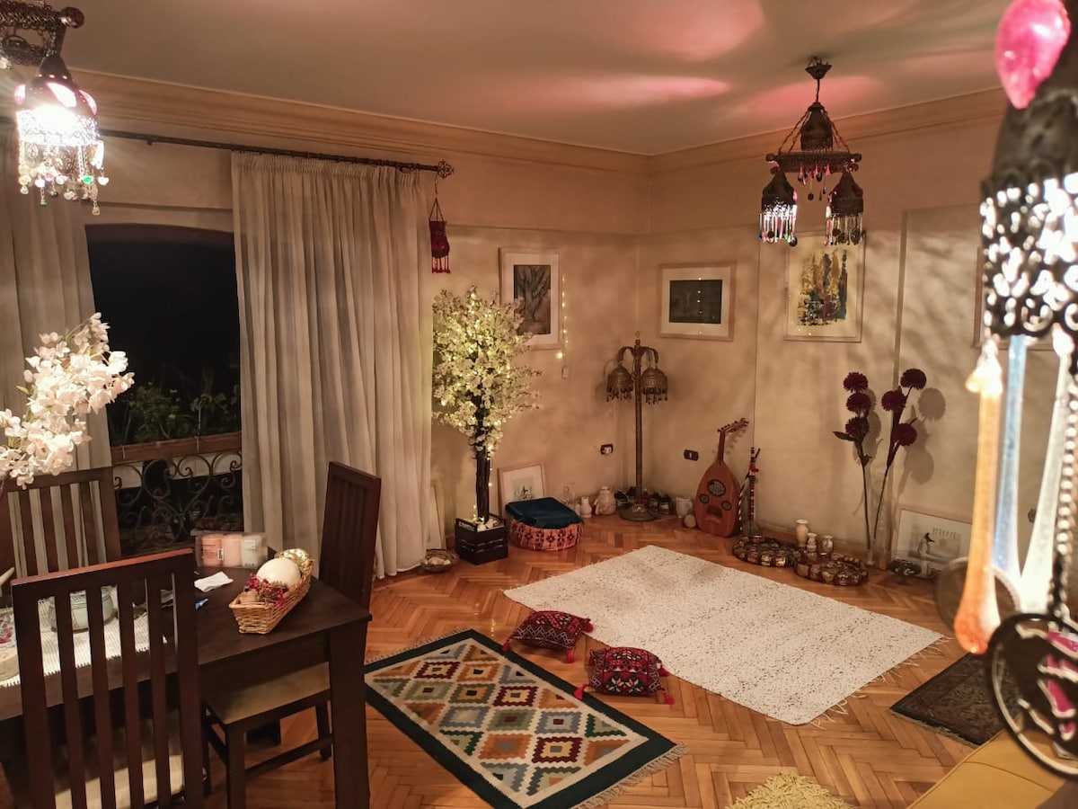 An inviting living area is highlighted by intricate hanging lanterns and decorative elements. A cozy dining table with wooden chairs sits beside a white carpet, surrounded by elegant plants and artwork adorning the walls, creating a serene atmosphere filled with cultural accents.