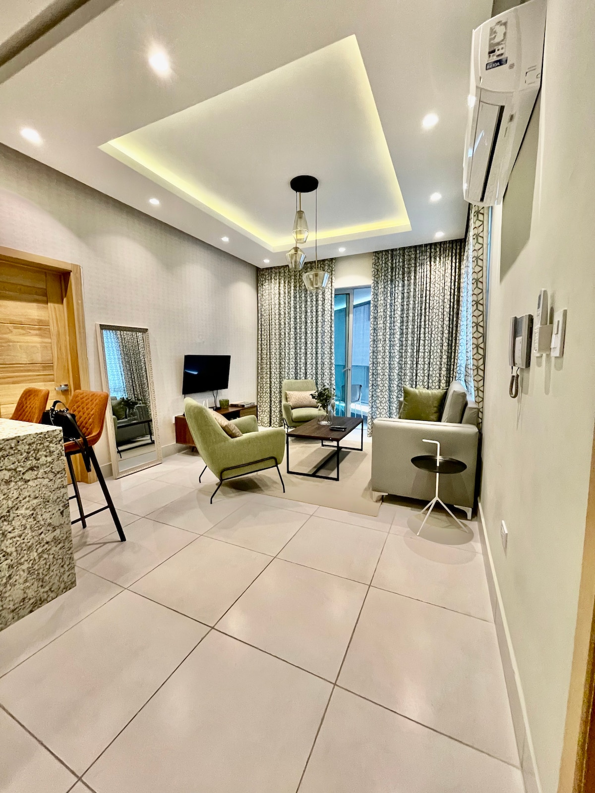 The living area features modern furnishings, including a combination of light and dark chairs around a central coffee table. A flat-screen television is mounted on the wall. Natural light is let in through large windows, adorned with patterned curtains, creating a bright and airy environment.