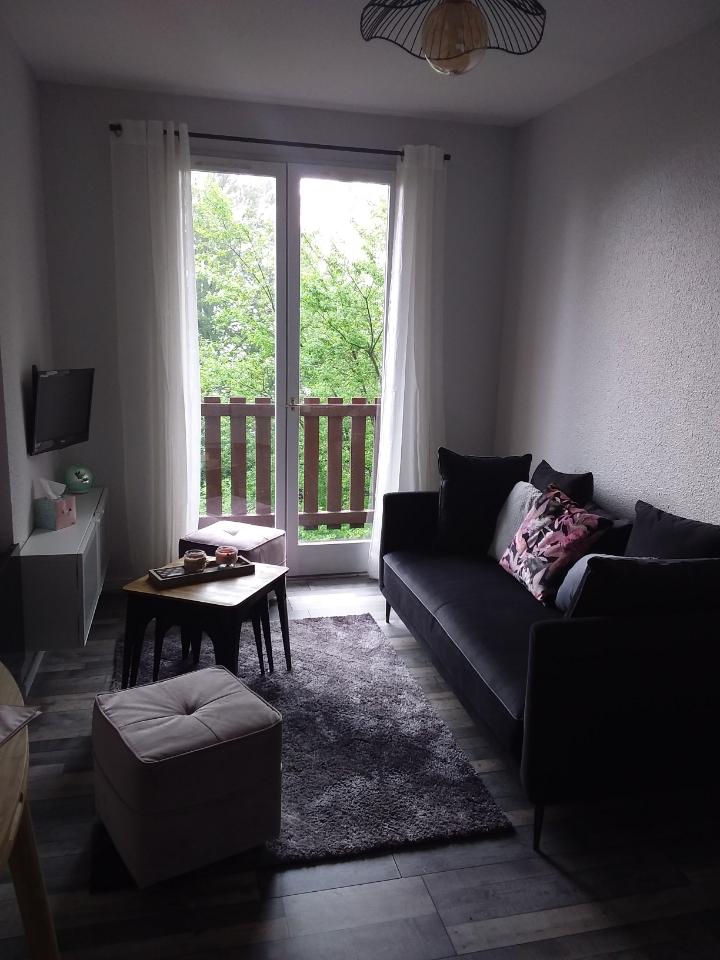 A cozy living area features a comfortable dark sofa adorned with decorative pillows. A small table is positioned in front, with magazines resting on it. Natural light enters through glass doors that open to a balcony surrounded by greenery.