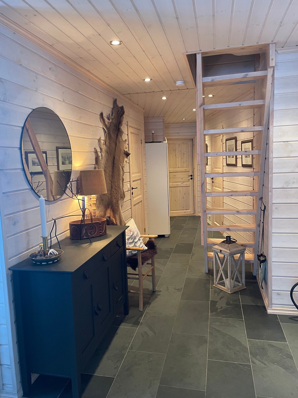 A spacious hallway featuring a light wooden interior and dark stone flooring. A circular mirror and lamp adorn a black console table, while a wooden chair and a decorative lantern are positioned nearby. A staircase leads to the upper level, and a refrigerator is visible in the background.