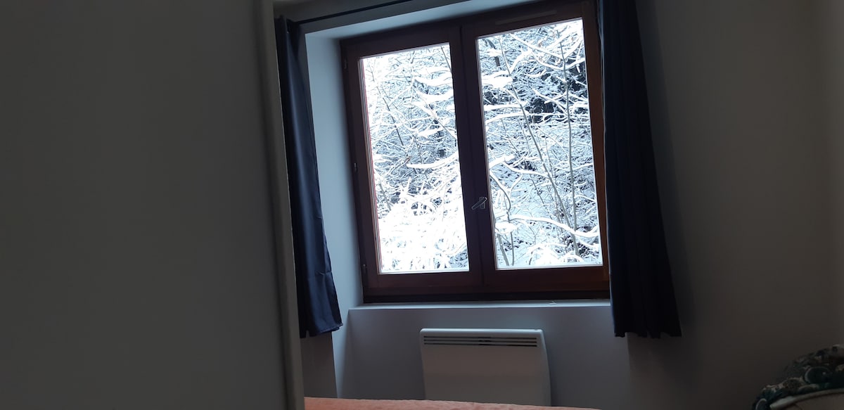 A window is framed by dark curtains, showcasing a serene snowy landscape outdoors. Frost-covered branches create a calming view, while the interior features a simple heating unit beneath the window, offering warmth during colder months.