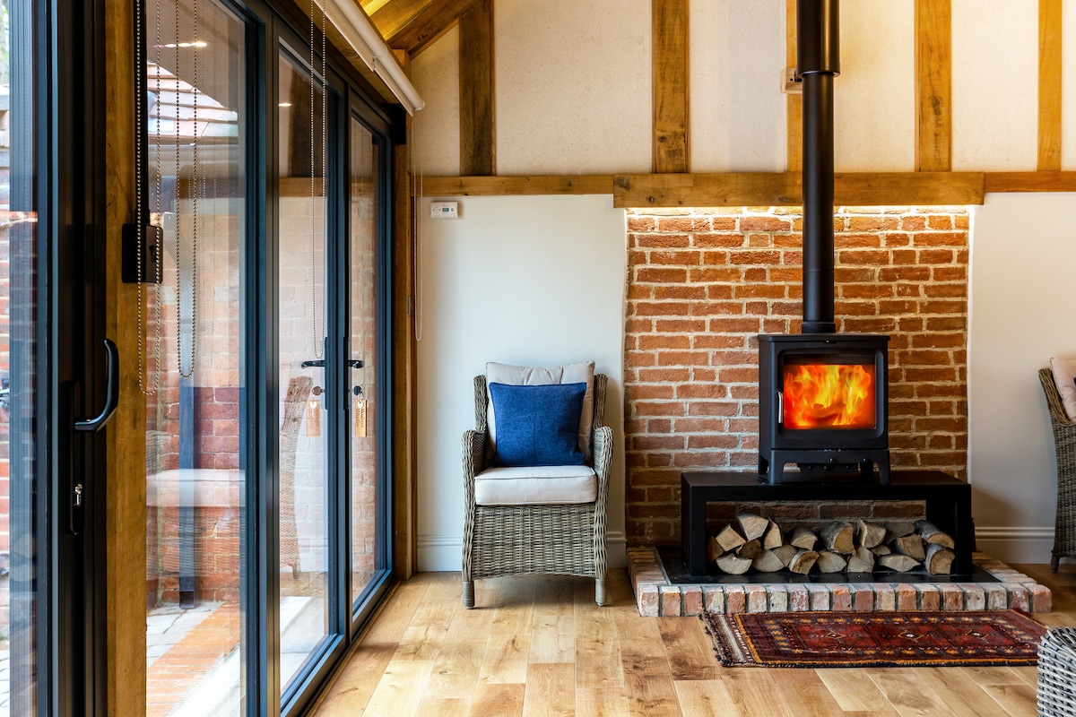 A cozy living area features a log burner set on a brick platform, surrounded by a wooden beam ceiling. Two wicker chairs, one with a blue cushion, are positioned near large glass doors that welcome natural light.
