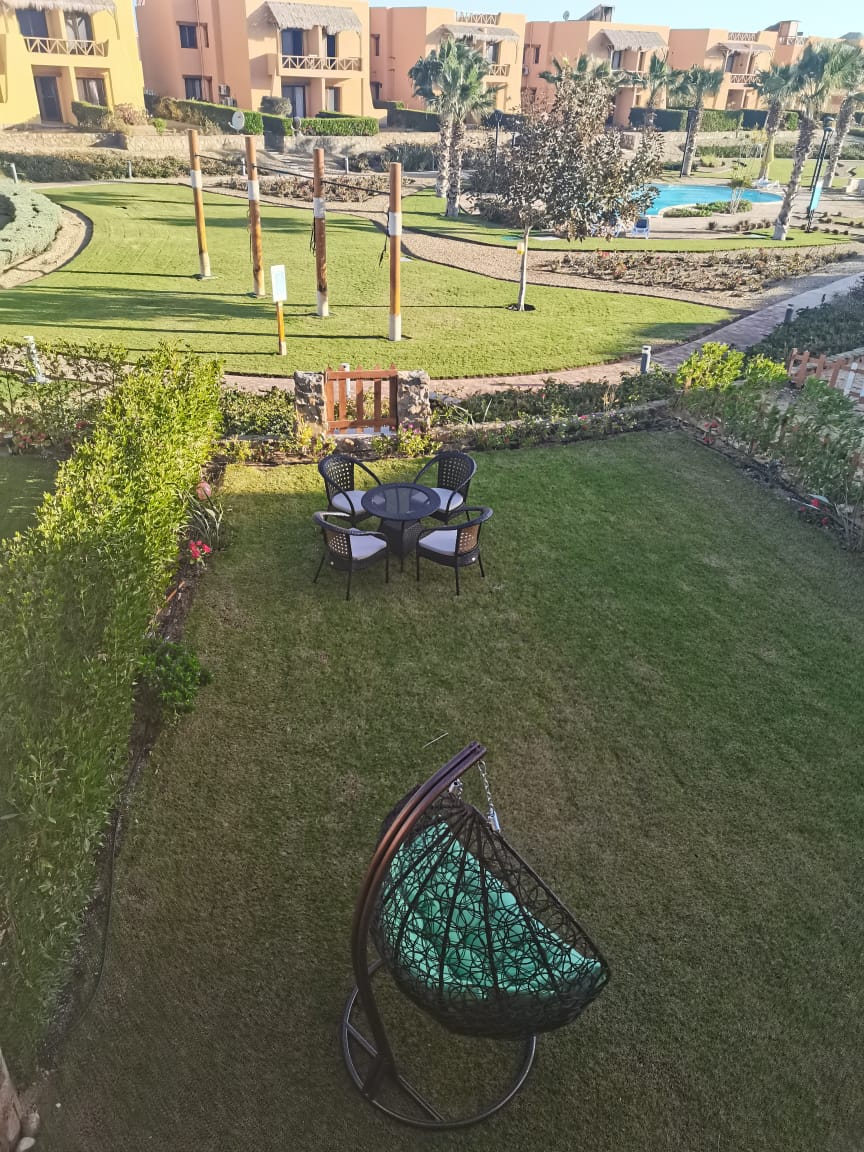 A well-maintained private garden features a circular table surrounded by four chairs, complemented by a hanging chair. Lush green grass and neatly trimmed hedges create a serene outdoor setting, providing views of the surrounding landscaped areas and nearby swimming pool.