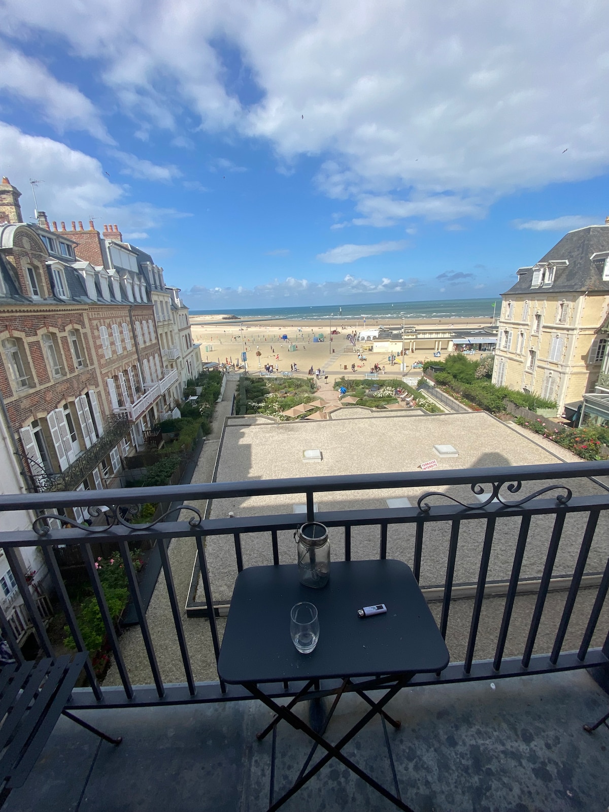 A balcony view is presented, featuring a small table and glassware. The scene overlooks a beach, where people can be seen enjoying the sandy shore. Elegant buildings line the street, complemented by a partly cloudy sky, adding depth to the coastal landscape.