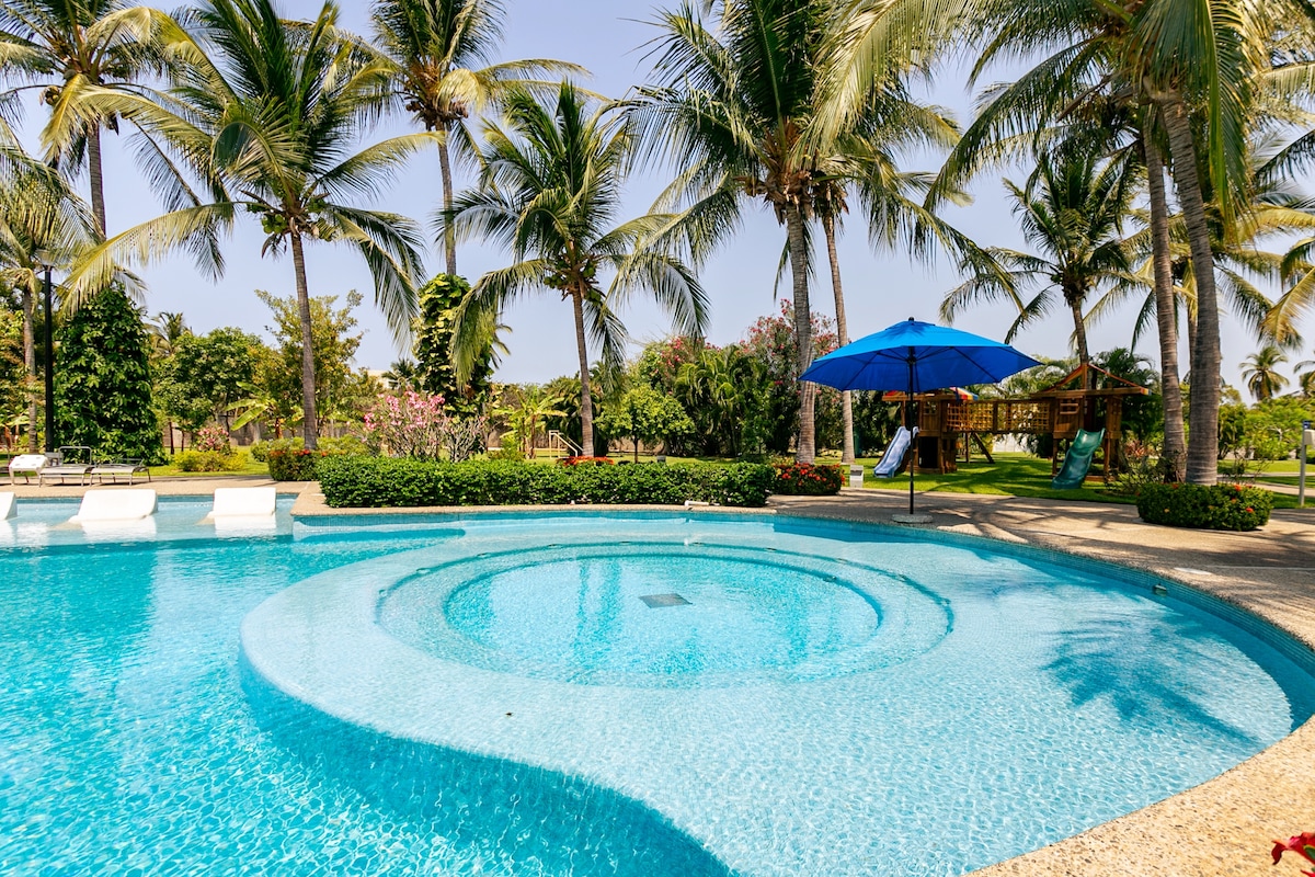 A swimming pool shaped with gentle curves is surrounded by lush palm trees and vibrant tropical landscaping. A blue umbrella provides shade over a seating area, while sun loungers are positioned nearby for relaxation. The clear water reflects the sunny sky, enhancing the serene environment.