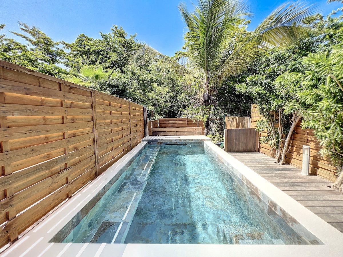 A private pool is showcased amidst lush greenery, enclosed by wooden fencing for privacy. Sunlight reflects on the clear water, creating a serene atmosphere. The surrounding area features wooden decking, enhancing the outdoor living space.