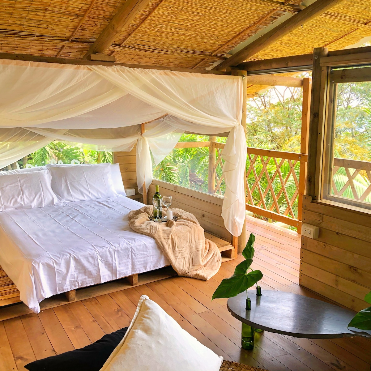 A spacious sleeping area features a double bed with white linens, draped with a sheer canopy. A soft blanket is casually placed on the bed. Natural light enters through large windows, enhancing the wooden interior and providing views of the surrounding greenery.