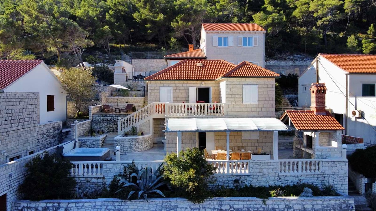 The villa's exterior showcases natural stone walls and a terracotta roof. A spacious terrace extends from the main house, furnished with outdoor seating. Lush greenery surrounds the property, enhancing the tranquil setting. Two hot tubs are visible on the terrace, inviting relaxation.