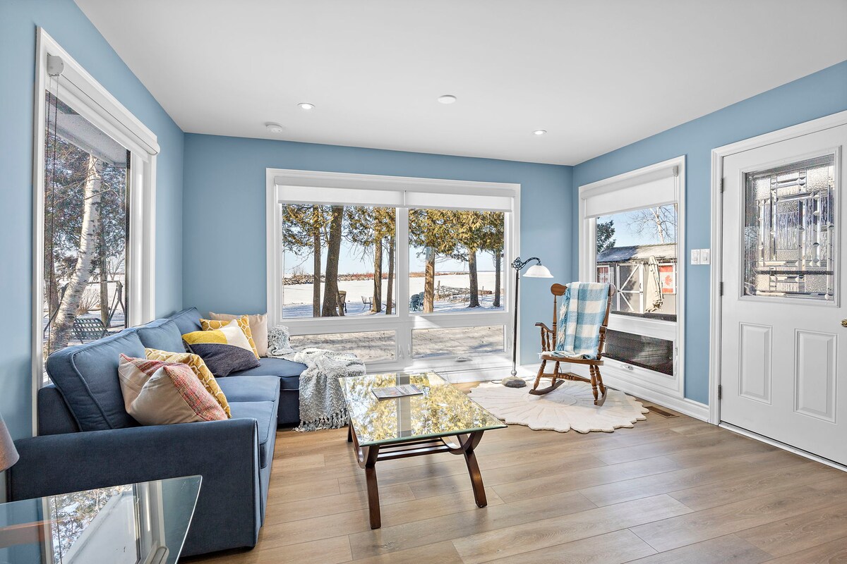 A bright living area on Lake Simcoe features large windows offering views of the water and trees. A comfortable blue sofa is complemented by decorative pillows. A glass coffee table is placed on a light wood floor, with a rocking chair near the side entrance.