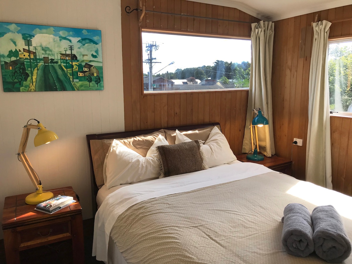 A well-lit bedroom features a spacious bed adorned with crisp white linens and a neutral-colored throw. Complimentary towels are neatly placed on the bed. A bedside table is positioned beside the bed, highlighted by a vintage-style lamp. Natural light filters through a large window with sheer curtains, illuminating the wooden accents.