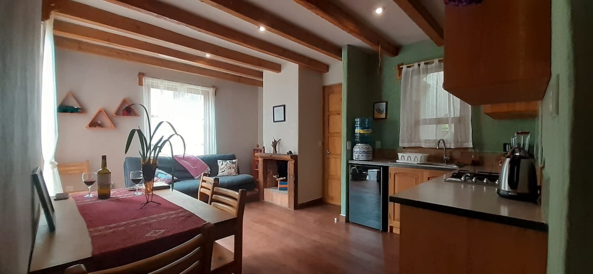 A warm living space showcases exposed wooden beams and a cozy seating area. A dining table is set for two, accompanied by a kitchenette featuring modern appliances. Natural light filters through sheer curtains, creating a bright and inviting ambiance.