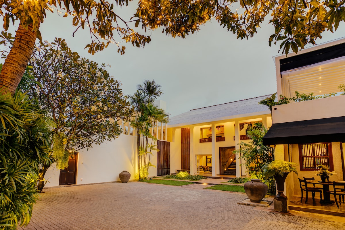An open courtyard is surrounded by lush greenery and decorative plants. A series of stone paths lead to a two-story building, highlighted by warm lighting from the upper floors. Decorative pots are positioned along the edges, creating a serene and welcoming environment.
