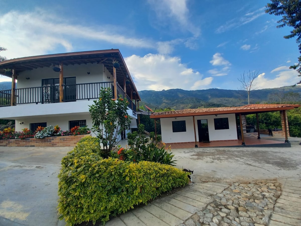 A spacious two-story house is situated against a backdrop of rolling hills. Flower beds with vibrant plants line the front, and a covered patio area is visible nearby, providing outdoor seating. The sky above features a blend of clouds and blue, adding to the serene landscape.