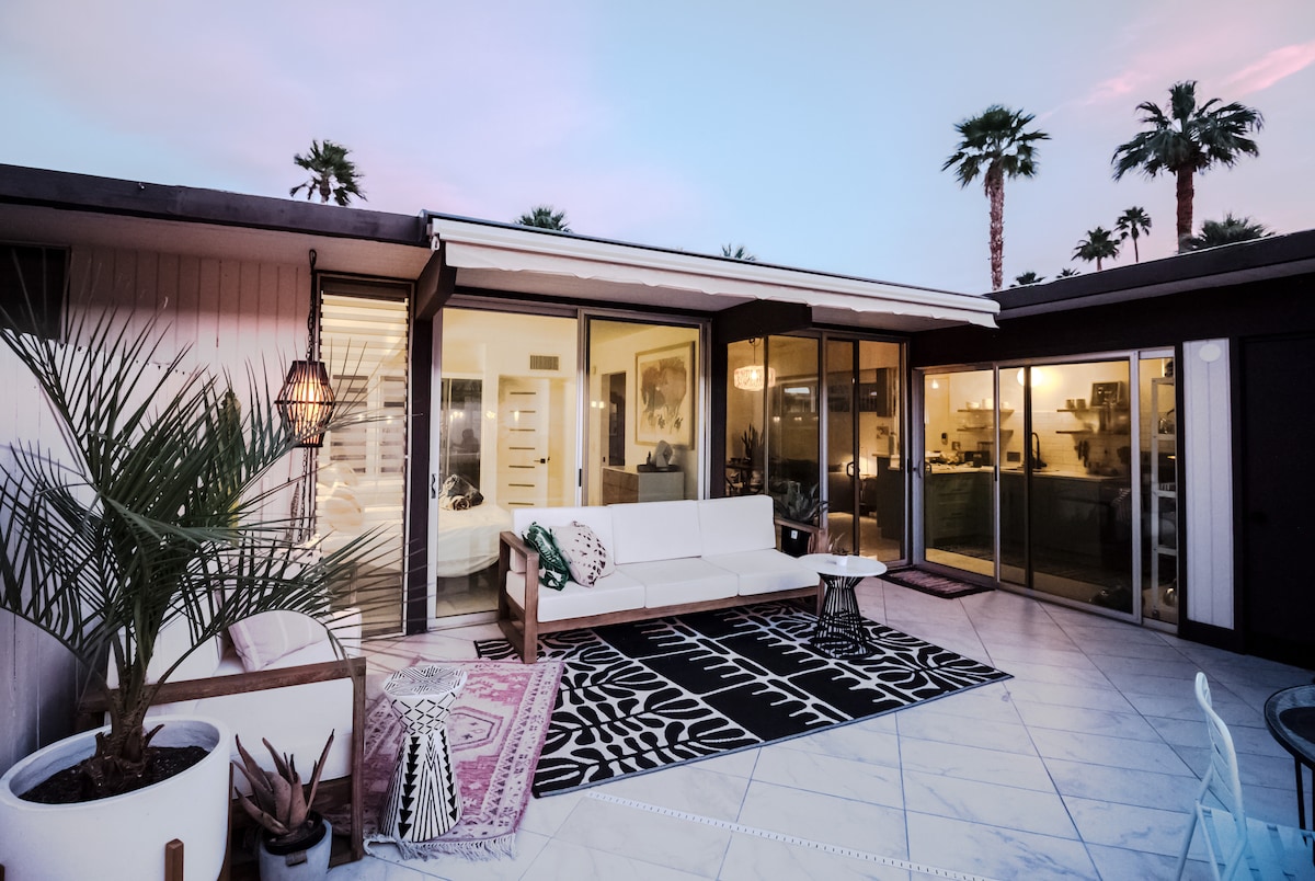 A private patio area features modern outdoor furniture, including a white sofa and accent table. Potted plants and a large rug add texture, while sliding glass doors provide access to the interior. Palm trees are visible against a twilight sky in the background.