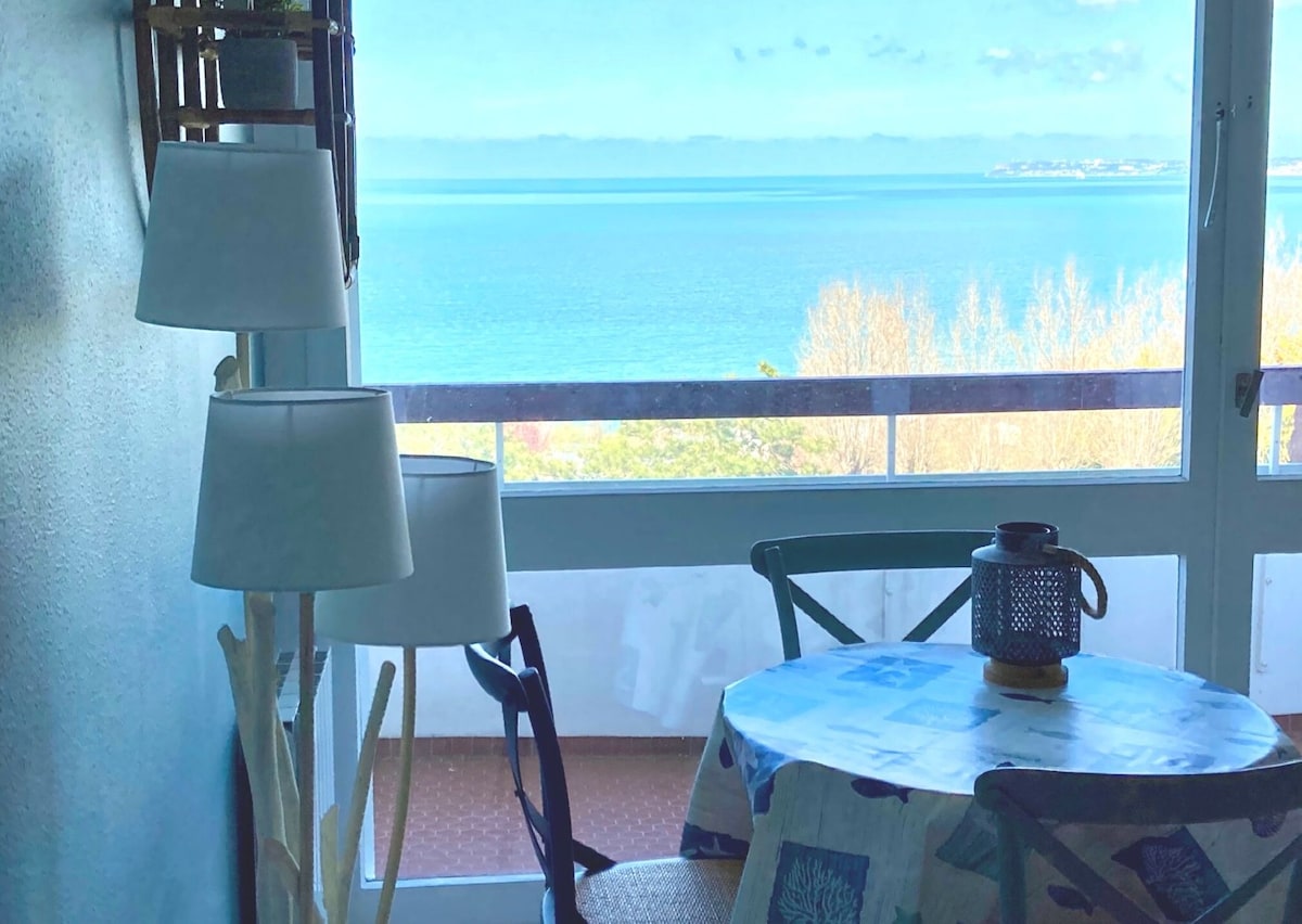A bright dining area features a round table with a patterned tablecloth, complemented by two chairs. A lamp and lantern provide soft lighting. Large glass doors open to a stunning view of the calm sea, enhancing the sense of serenity.