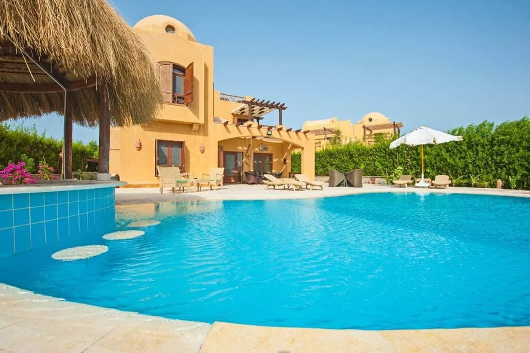 A bright pool area features a spacious layout highlighted by a turquoise swimming pool. Sun loungers are positioned on the surrounding deck, and a shaded area is provided by a thatched roof. Lush greenery lines the background, creating a serene outdoor environment.