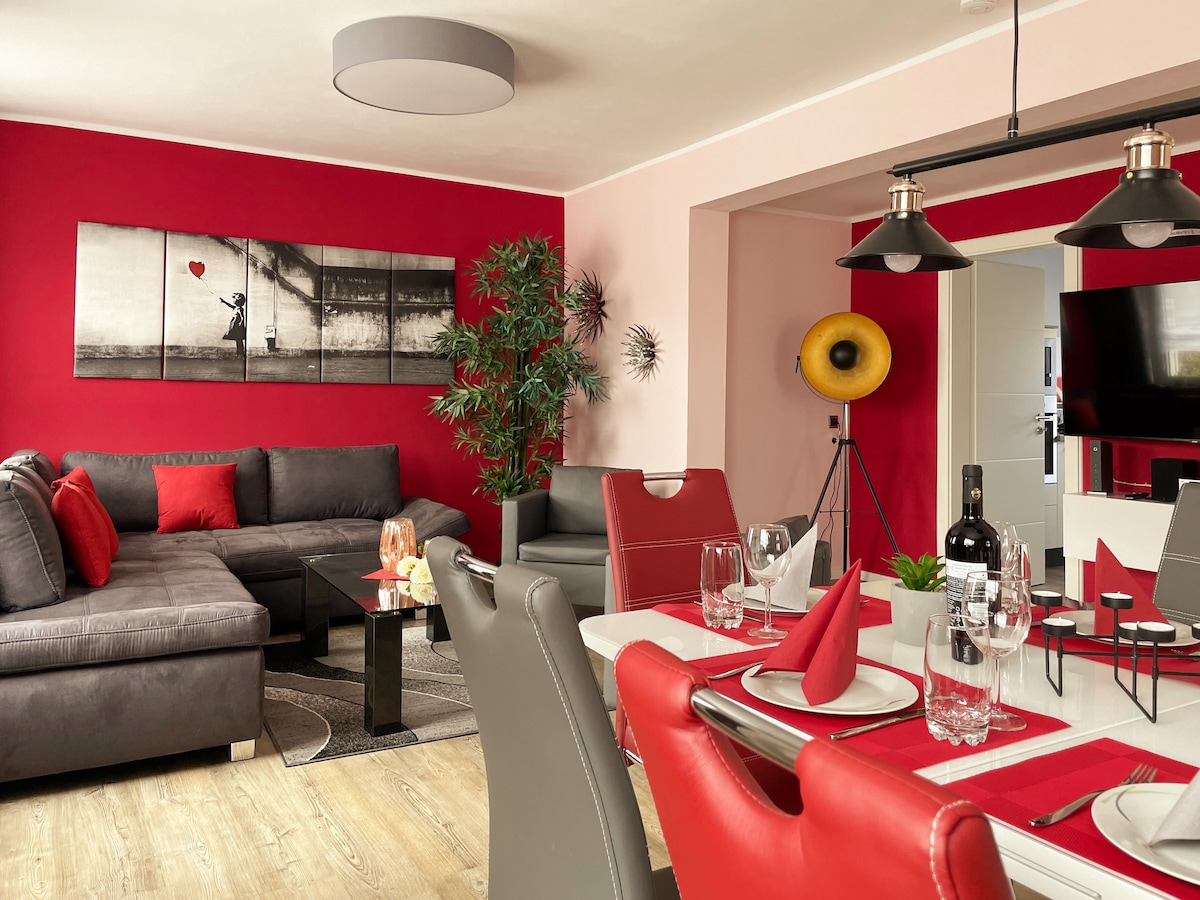 A spacious living and dining area features a sectional gray sofa and a sleek dining table set for six. Bold red walls provide a vibrant backdrop. Modern decor includes a large wall art piece and a standing lamp. A 55-inch Smart TV is visible in the corner.