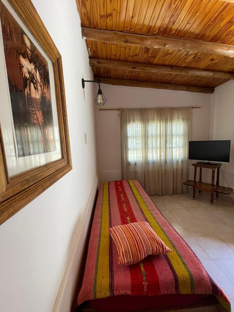 A bright room features a single bed adorned with a multi-striped blanket and a decorative pillow. A framed artwork hangs on the wall, and a small wooden TV stand supports a television. Natural light filters through sheer curtains, illuminating the space.
