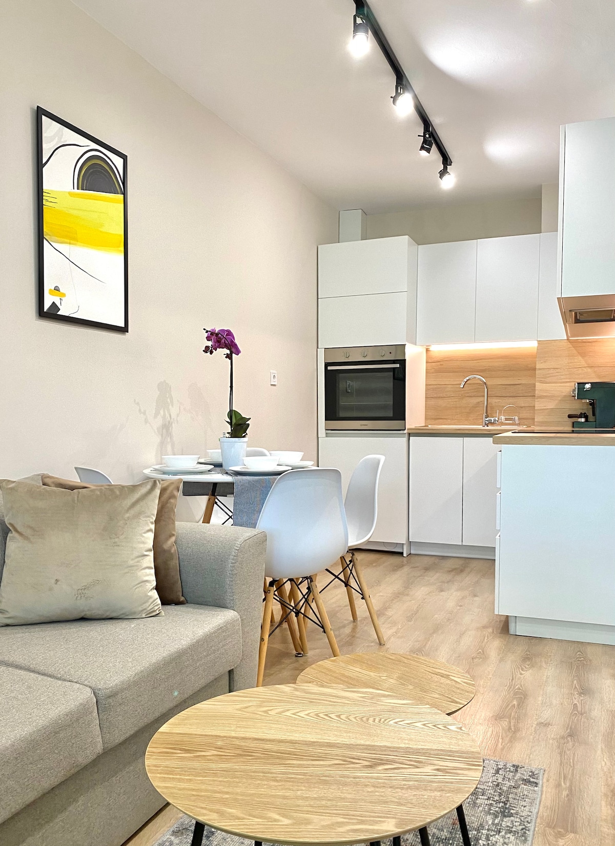 An open-plan kitchen and living area is featured with a modern design. A small dining table with four chairs is situated beside the kitchen, which includes sleek cabinets and appliances. A comfortable gray sofa and two wooden coffee tables are positioned within the space, complemented by decorative elements.