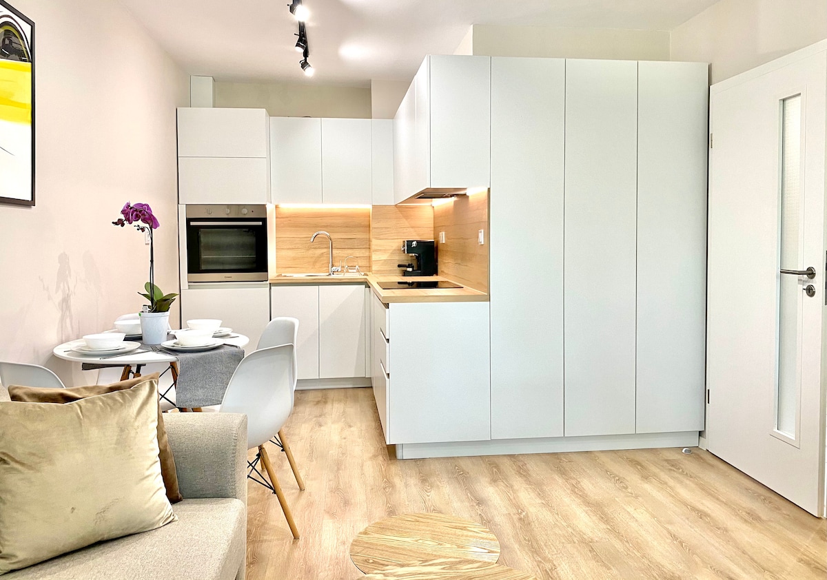 A modern kitchenette features sleek white cabinetry and wooden accents. A compact dining table with light grey chairs is set for two, accompanied by a small round coffee table. Natural light highlights the clean lines and functionality of the space.