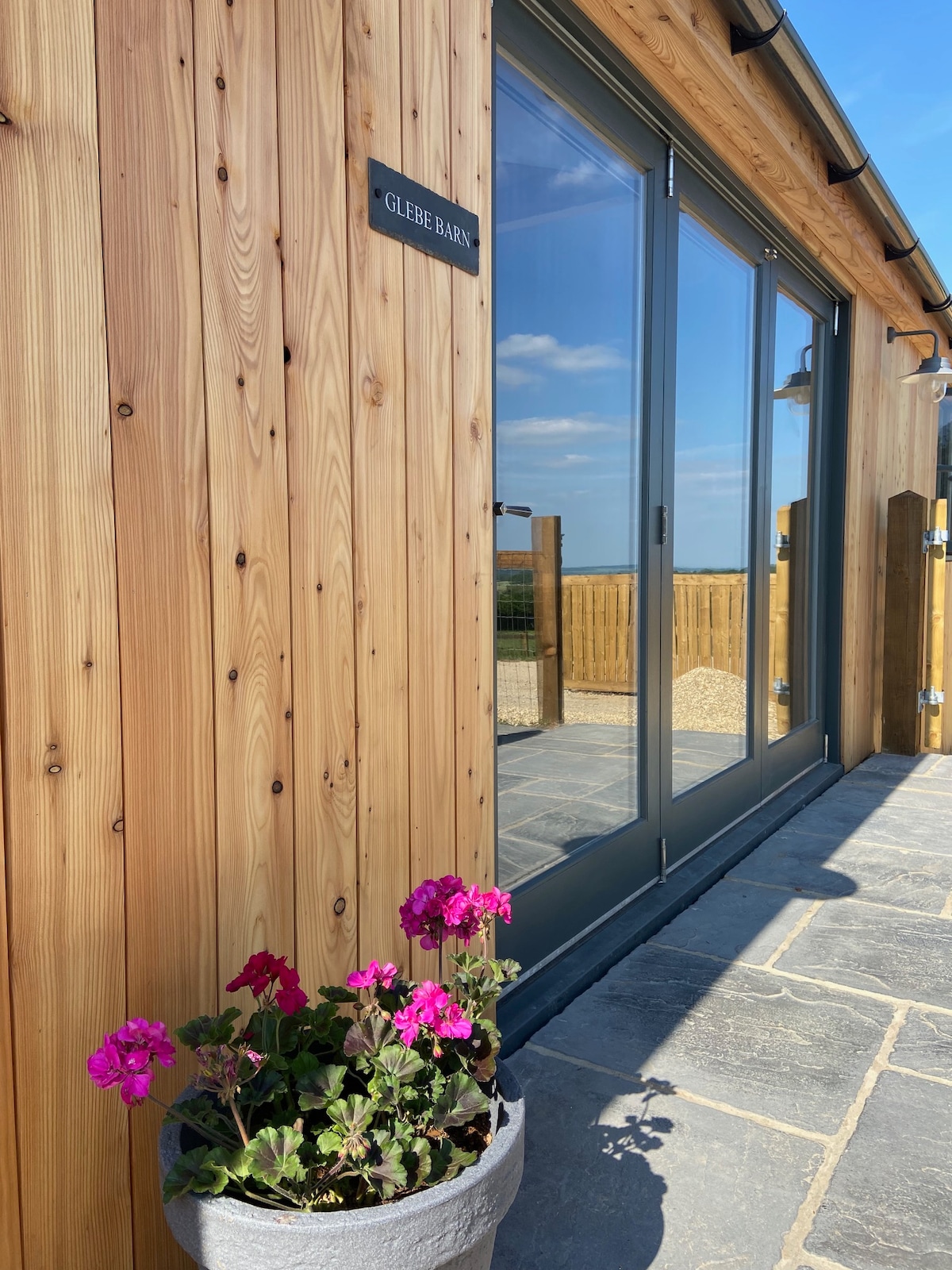 The exterior of Glebe Barn features a warm wooden facade with a clear view of large glass doors leading to the terrace. A potted plant with vibrant pink flowers adds a touch of colour to the stone pathway.