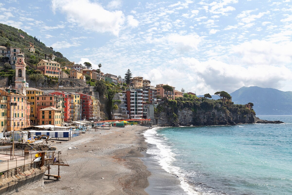 A coastal view is presented, featuring a sandy beach alongside a clear blue sea. Colorful buildings line the shore, with a backdrop of lush hills. The sky is partly cloudy, adding to the serene atmosphere of the seaside village.