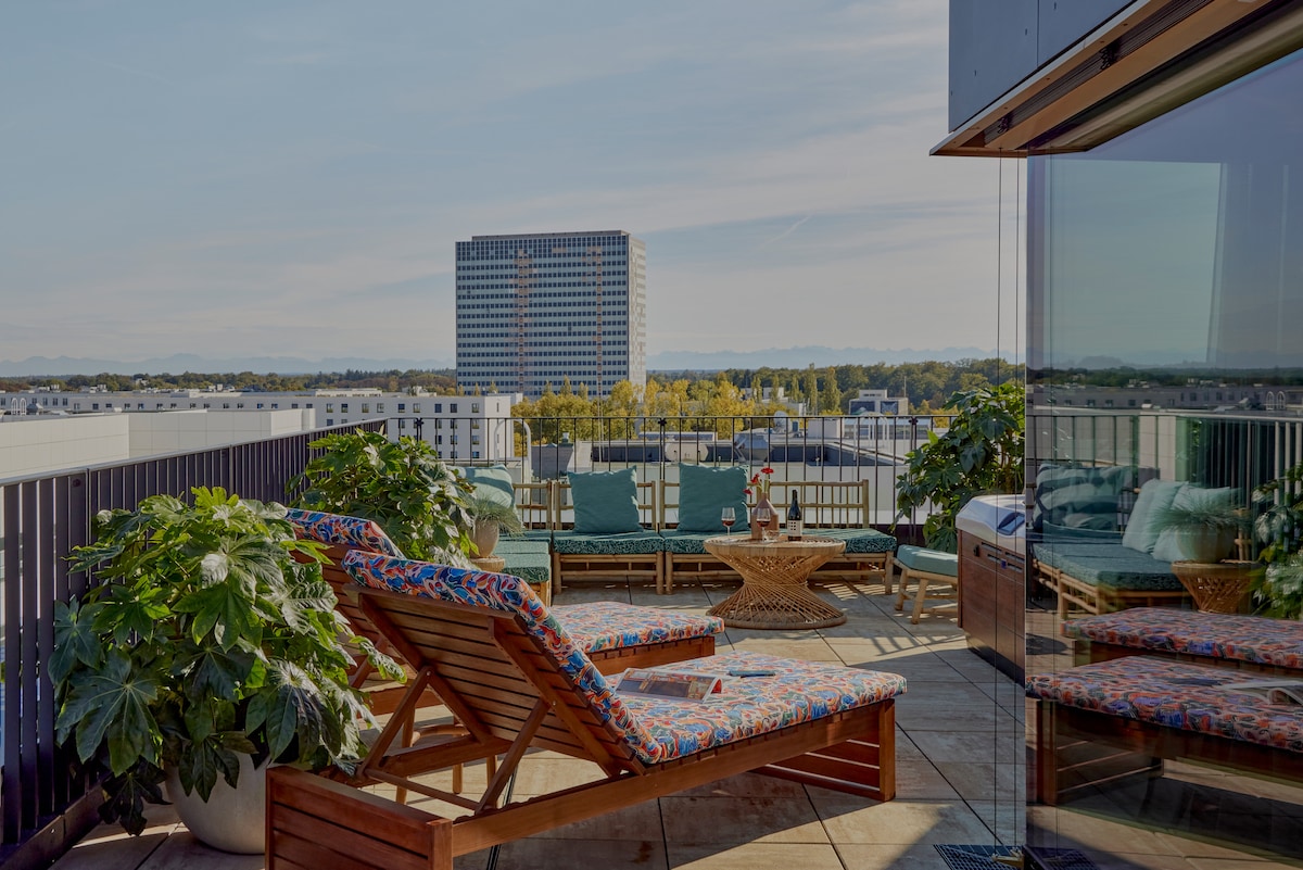 A spacious rooftop terrace features vibrant lounge chairs and a natural wood table, surrounded by lush greenery. A panoramic view includes a distant building and a clear blue sky, creating a serene outdoor setting for relaxation.