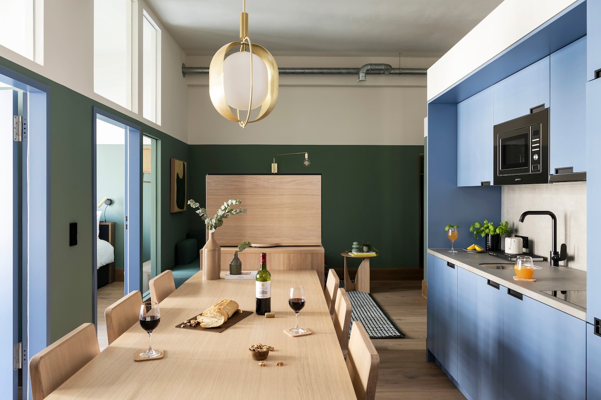 A spacious dining area features a large wooden table surrounded by beige chairs, creating an inviting space for gatherings. The kitchen, equipped with modern appliances and a microwave, showcases blue cabinetry. Decorative touches include a vase of flowers and a bottle of wine on the table.