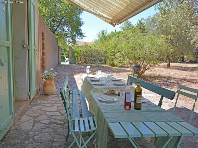 An outdoor dining area is set on a stone patio, featuring a green tablecloth draped over a table surrounded by metal folding chairs. Glasses and bottles are arranged on the table, with greenery and trees visible in the background. A BBQ grill is positioned nearby.