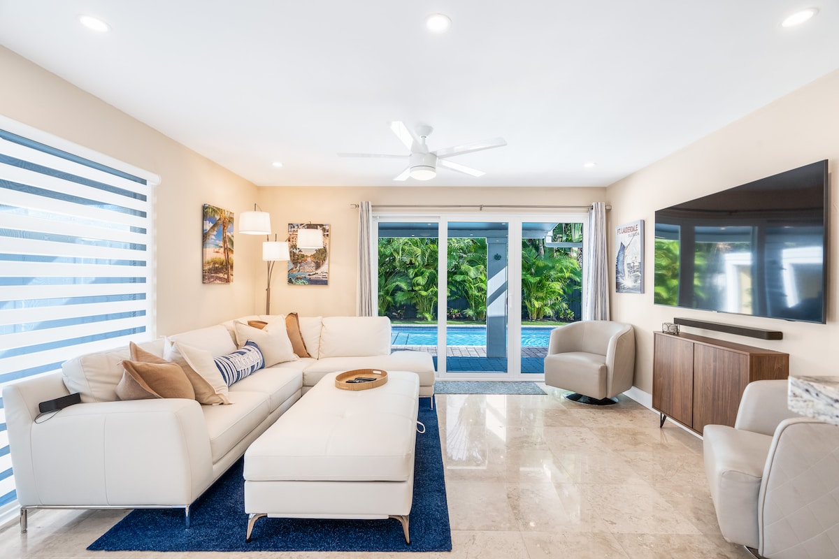 A light-filled living room features a spacious sectional sofa and stylish accent chairs. Large glass doors open to the pool view, enhancing the airy atmosphere. Tropical artwork adorns the walls, and a sleek media console holds a television, creating a comfortable relaxation space.