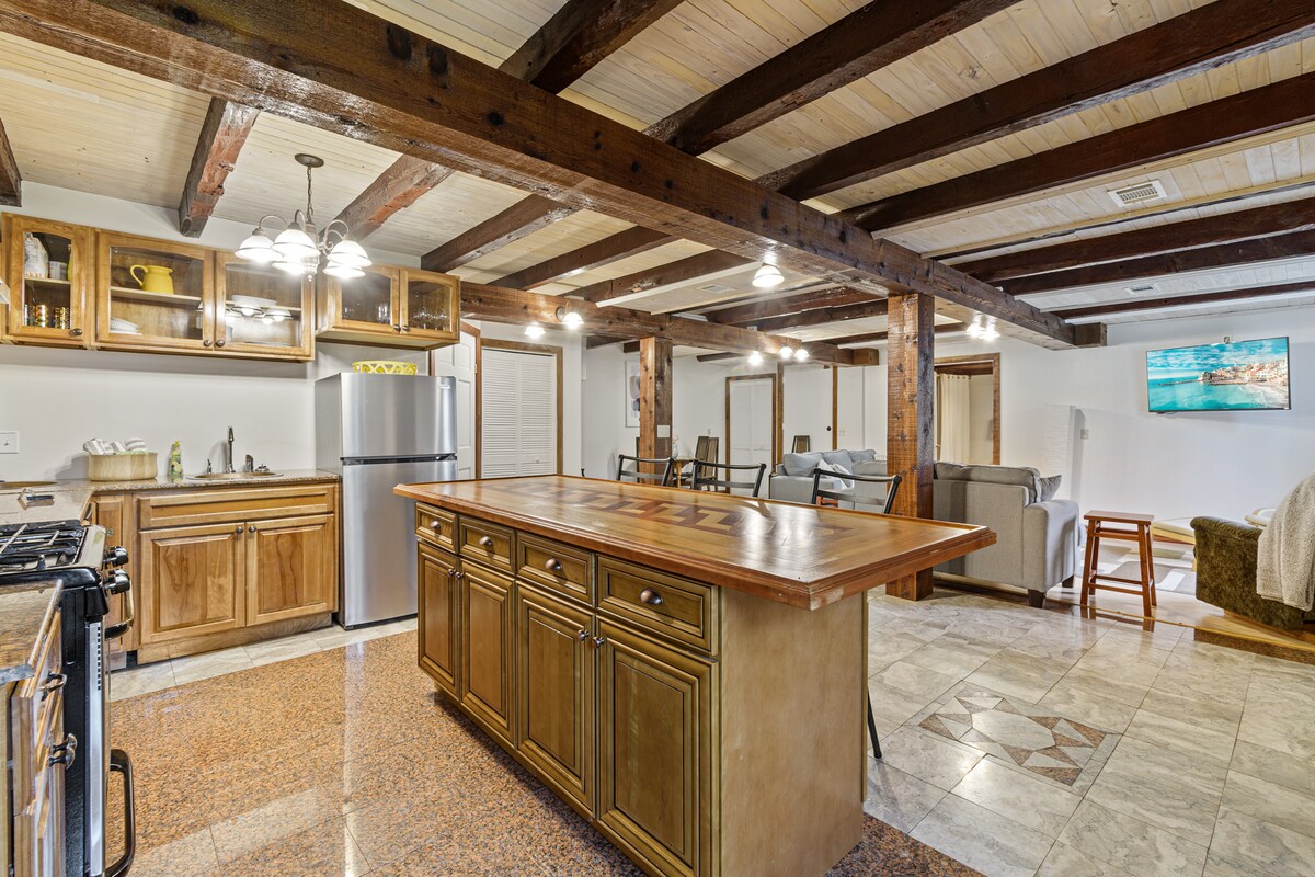 An open kitchen and living area features exposed wooden beams and a large central island with a polished countertop. Modern appliances are visible, including a stainless steel refrigerator. The living area contains comfortable seating and a television positioned against the wall.