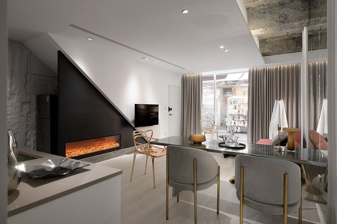 A modern living area is presented with a sleek fireplace along one wall and a flat-screen TV mounted above. Elegant furniture includes minimalist armchairs and a dining table. Natural light fills the room through large windows, complemented by soft drapes.