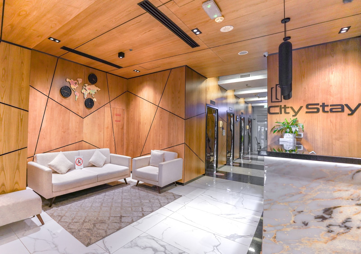A modern lobby area features natural wood panels on the walls, with a plush seating arrangement consisting of two light-colored sofas. A patterned rug is placed on a marble floor, and a decorative plant is seen in the corner, creating a welcoming entrance.