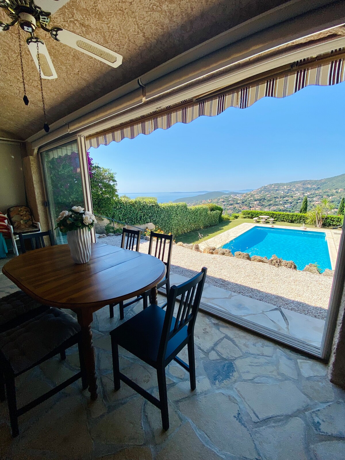 A dining area is presented with a wooden table surrounded by black chairs. The large glass doors offer an unobstructed view of the pool and the hills, with lush greenery visible in the background. Natural light floods the space, enhancing the inviting atmosphere.