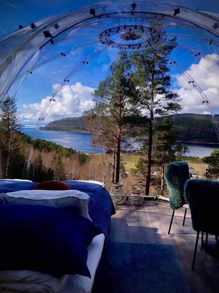 A clear dome structure offers panoramic views of the surrounding landscape, featuring a serene lake and lush greenery. Inside, a spacious bed is adorned with soft linens, while two inviting chairs provide a cozy seating area. Natural light floods the space through the dome's transparent walls.