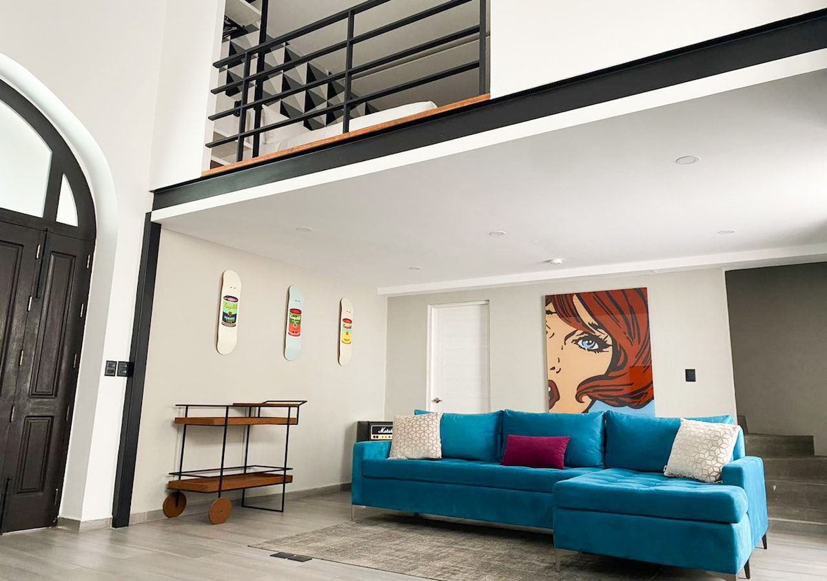 A spacious living area is presented with a vibrant blue sofa and a central coffee table. Art featuring a retro portrait adorns the wall, alongside decorative skateboards. A staircase leads to a lofted sleeping area, while a door and windows allow natural light to brighten the room.