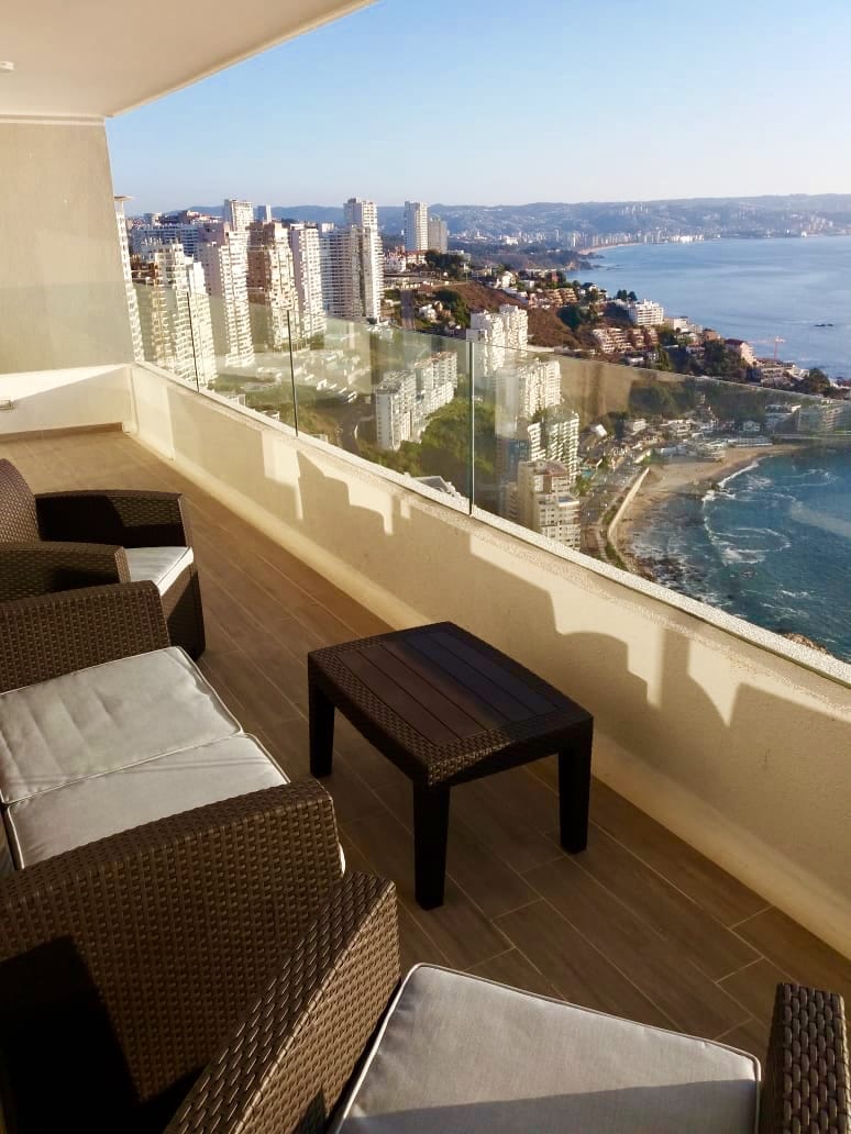 A spacious terrace is seen, featuring a comfortable seating area with several wicker chairs and a small table. The backdrop includes a stunning view of the coastline and cityscape, with glass railings providing unobstructed sightlines of the ocean and surrounding landscape.