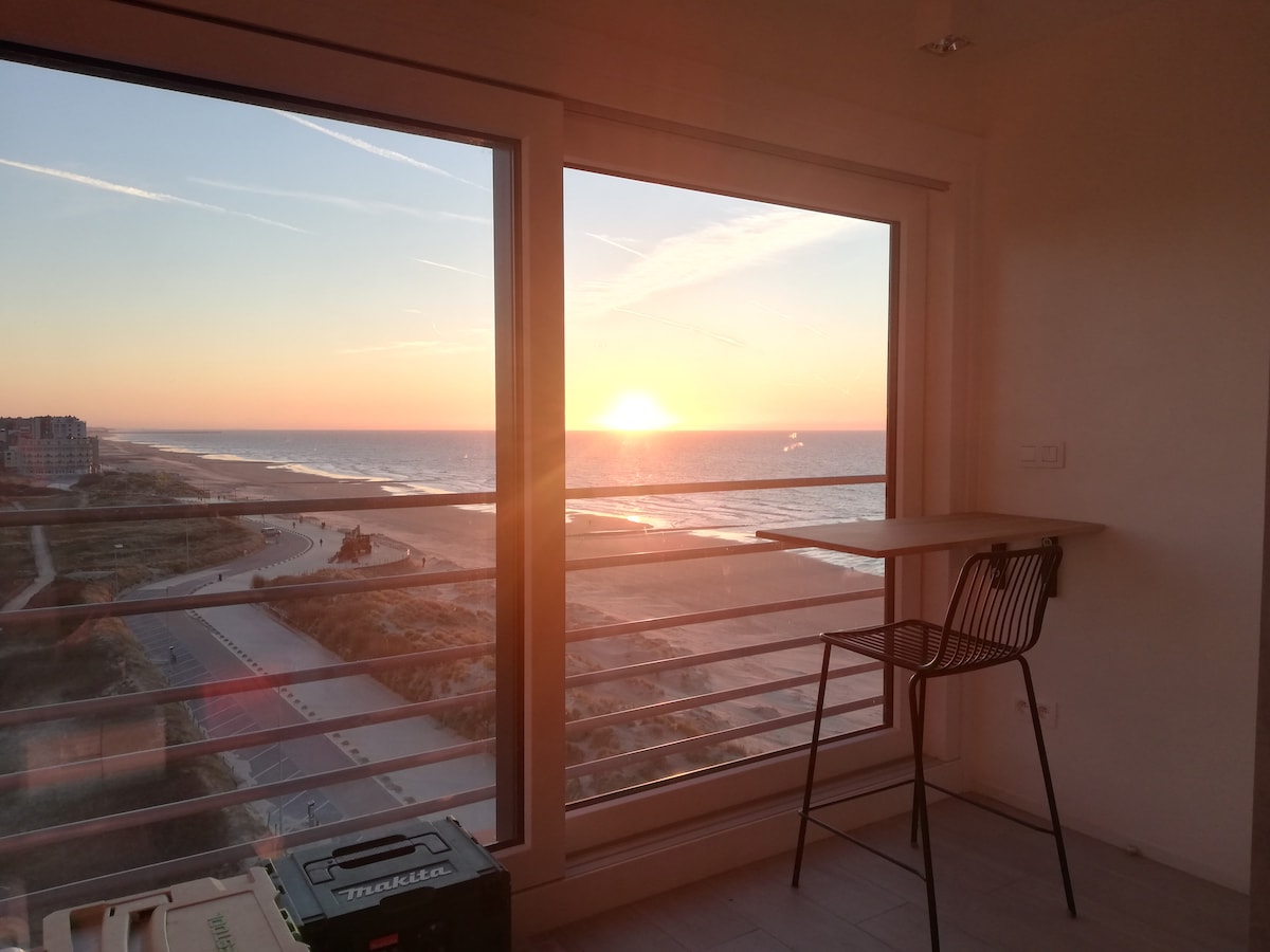 A stunning view of the sunset over the ocean is captured through floor-to-ceiling windows. A small side table and a chair are positioned nearby, providing a spot to enjoy the scenery. The beach and promenade are visible in the foreground, framed by soft evening light.