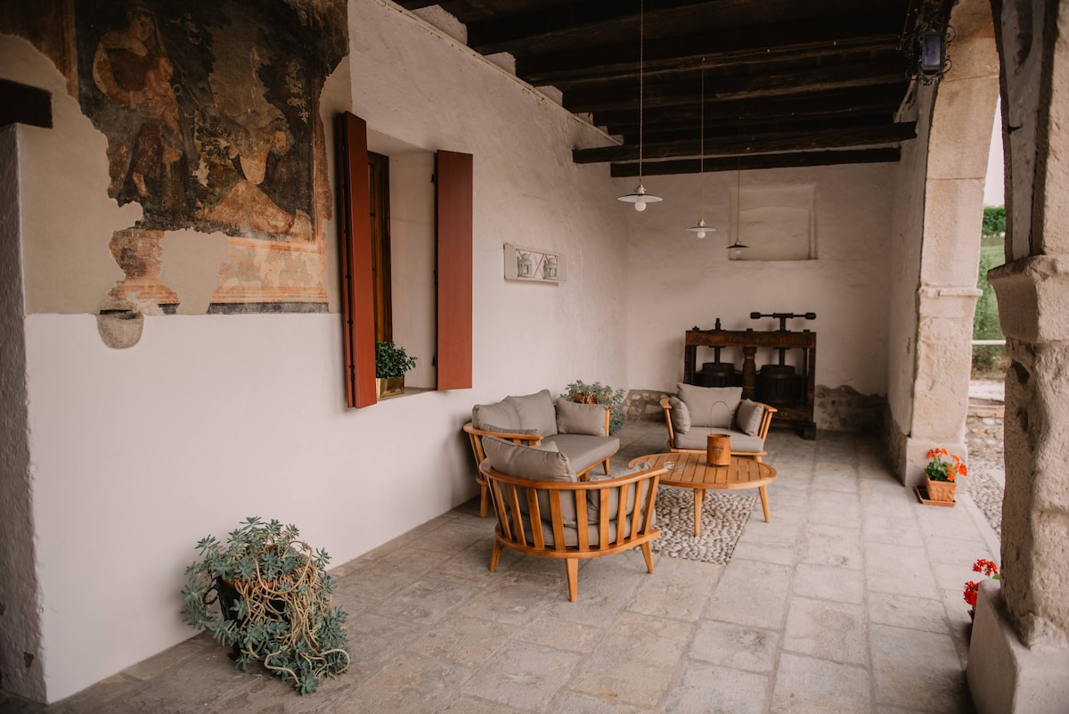 A shaded porch area features comfortable wooden seating arranged around a small table. Decorative plants add greenery, while wall murals hint at the historic character of the space. Soft lighting fixtures are suspended from the ceiling, enhancing the relaxed atmosphere.