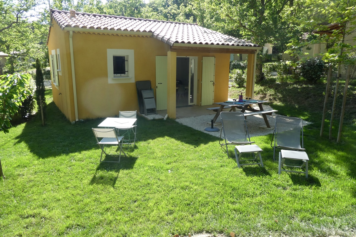 A charming yellow cottage is surrounded by a lush green lawn. Outdoor furniture includes a wooden table with benches, four lounge chairs, and a covered terrace. The property is framed by trees that provide a serene, natural setting.