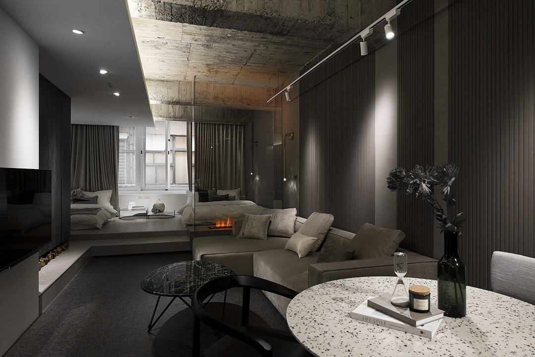 A modern living space features a cozy seating area with a sectional sofa and a coffee table topped with decorative items. Natural light fills the room through large windows, and a sleek bed is visible in the background. Textured walls and a concrete ceiling add an industrial touch.