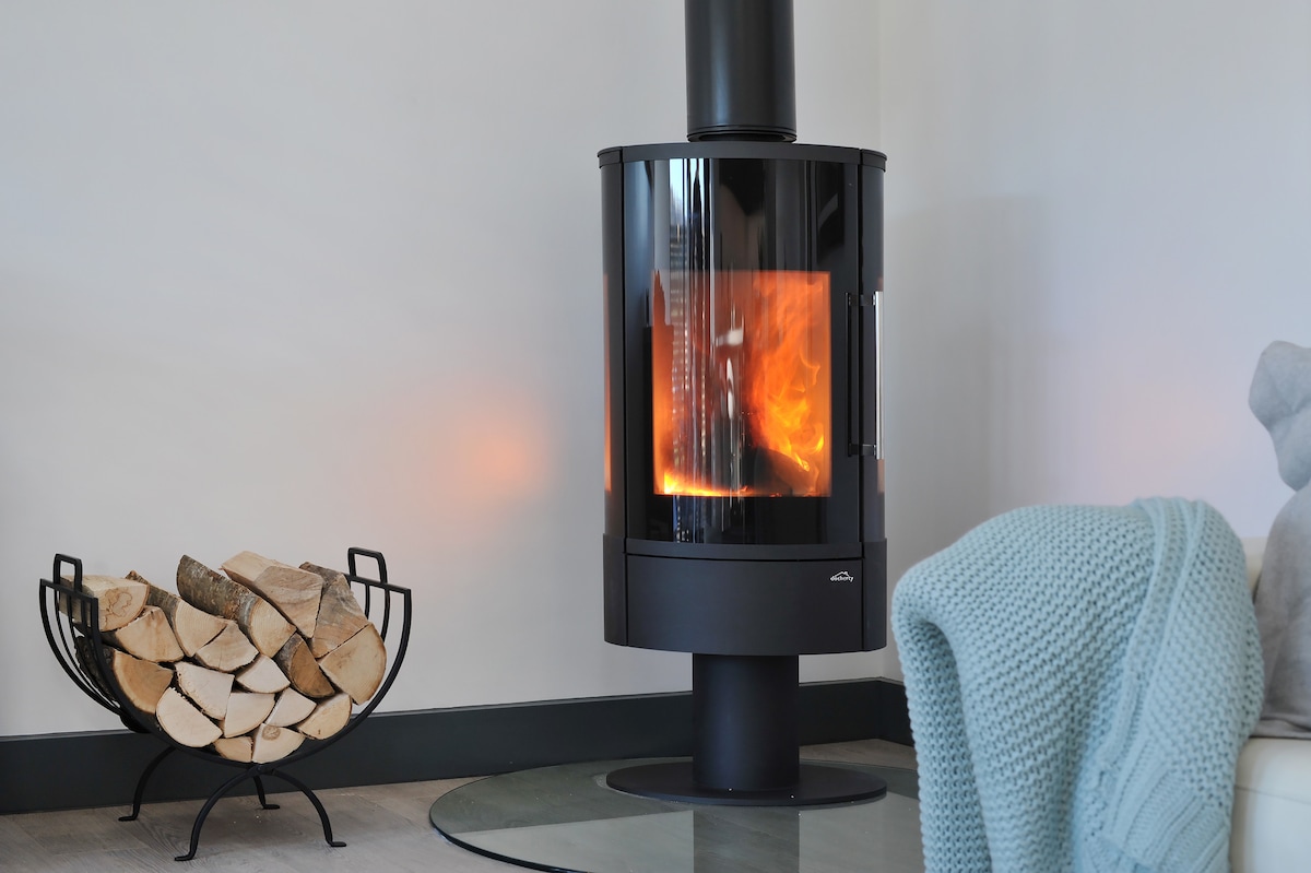 A contemporary wood-burning stove stands at the center of the living area, featuring glass panels that showcase a warm, flickering flame. Beside it, a stylishly arranged basket holds neatly stacked logs, enhancing the inviting ambiance.